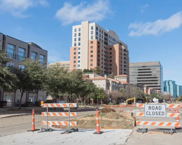Road closed sign in Downtown Irving, Texas, USA under cloud blue sky. Barricade closures, cones with construction equipments and high-rise building in background.