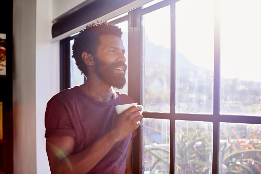 Shot of a cheerful young man standing next to a window and looking through it while drinking a cup of coffee inside at home during the day