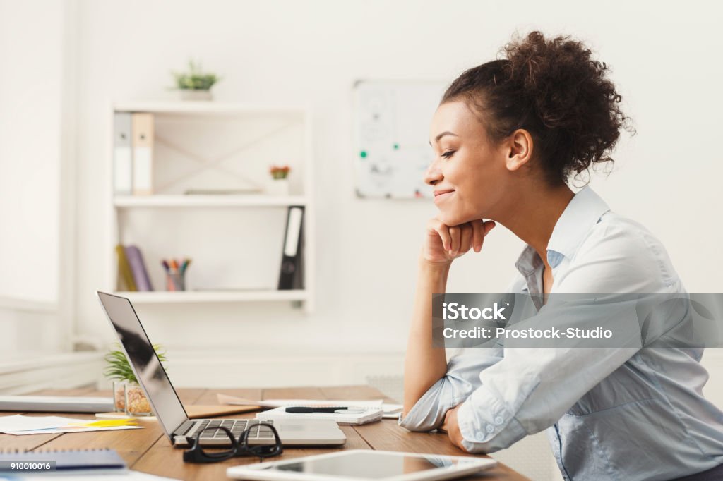 Happy business woman working on laptop at office Happy smiling african-american business woman working on laptop at office. Businesswoman sitting at her working place, copy space, side view Benefits Stock Photo