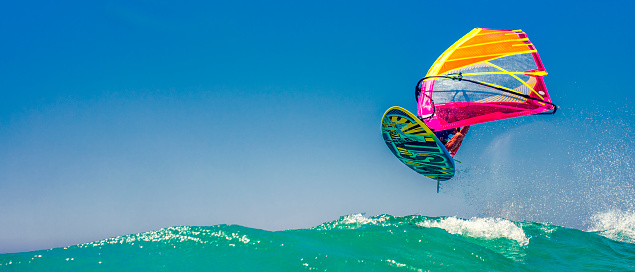 Photo of a windsurfer in mid-air during a jump over a wave in a turquoise sea.