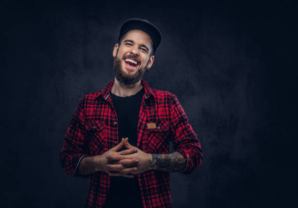 A bearded stylish hipster male with a tattoo. A caucasian bearded hipster male laughing dressed in a flannel shirt and a cap, posing in studio, over dark background. man beard plaid shirt stock pictures, royalty-free photos & images