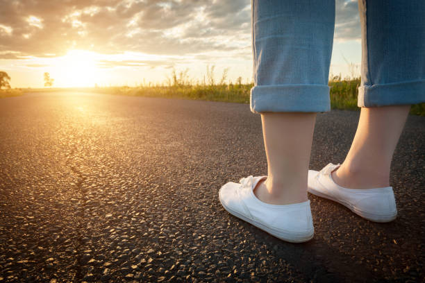 太陽に向かってアスファルト道路に立っている白いスニーカーの女性。旅行、自由の概念。 - foot long ストックフォトと画像