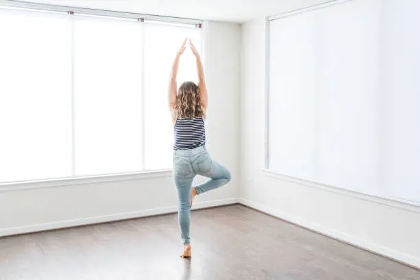 Young woman in tree yoga pose standing on one leg in empty modern new room, hardwood floors, large sunny windows in apartment, house