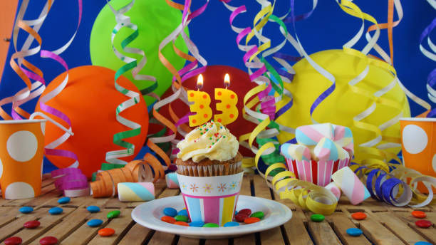 Birthday 33 cupcake with candles burning on rustic wooden table with background of colorful balloons, plastic cups and candies with blue wall in the background Cupcake in the center of the image with colorful candles burning on rustic wooden table with background of colorful balloons, streamers, plastic cups and candies with blue wall in the background number 33 stock pictures, royalty-free photos & images