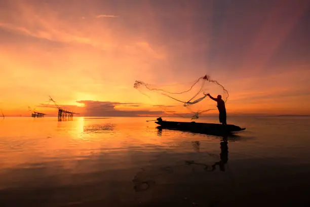 Photo of Asian fisherman on wooden boat casting a net for catching freshwater fish in nature river in the early morning before sunrise