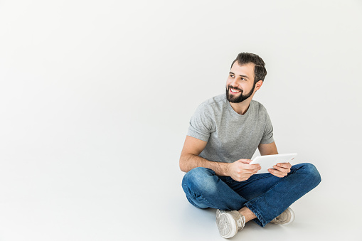 smiling bearded man using digital tablet and looking away while sitting isolated on white