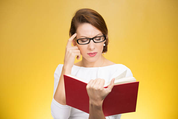 Young elegant woman in eyeglasses having problems with understanding while reading book isolated on yellow background