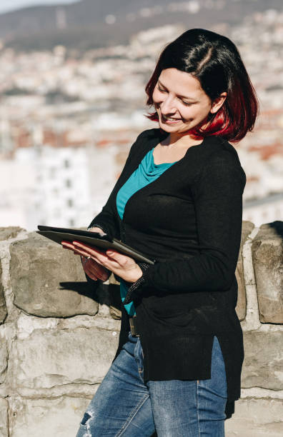 istock Happy smilling handsome redhead young woman sitting on a small wall working on tablet - Concept Relaxing in nature. 910156502