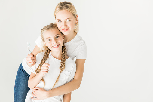 happy mother and daughter holding toothbrushes and smiling at camera isolated on white