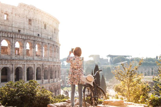 schöne junge frau in bunte mode kleid allein auf einem hügel mit fahrrad nimmt bilder des kolosseums in rom mit smartphone-kamera bei sonnenuntergang. touristisch attraktiven mädchen mit eleganten strohhut - journey elegance people traveling architecture stock-fotos und bilder