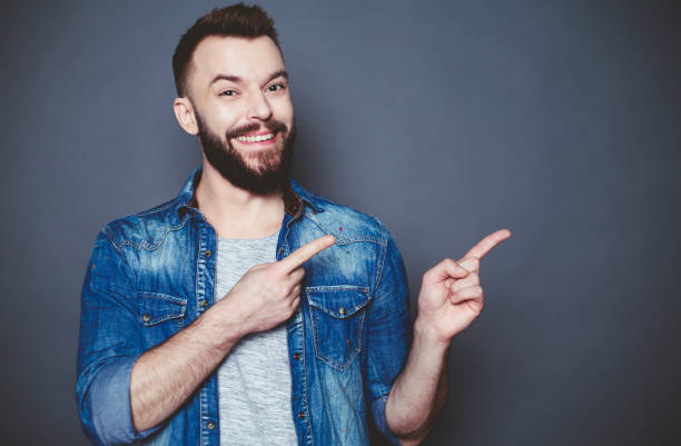istock Handsome smiling modern man in a denim shirt points his fingers away from himself on a gray background. 910170118