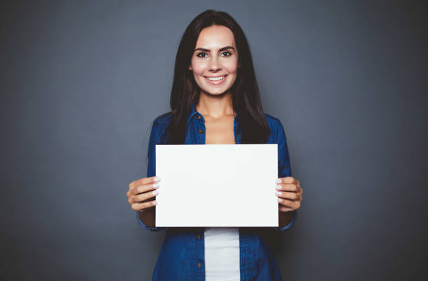 bella mujer sonriente moderno en una camisa de jeans con hoja en blanco de papel para publicidad en las manos sobre un fondo gris aislado. - paper greeting card blank document fotografías e imágenes de stock