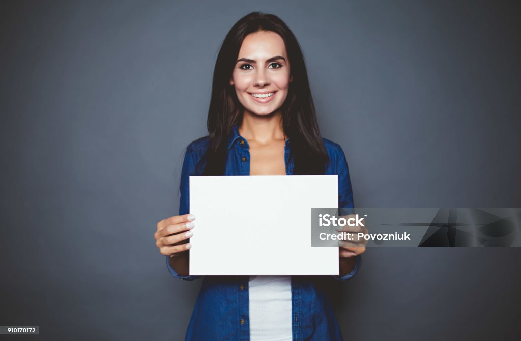 Beautiful modern smiling woman in a jeans shirt with blank sheet of paper for advertising in hands on a gray background isolated. Holding Stock Photo