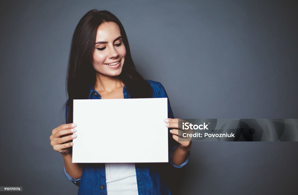 Bella mujer sonriente moderno en una camisa de jeans con hoja en blanco de papel para publicidad en las manos sobre un fondo gris aislado. - Foto de stock de Agarrar libre de derechos