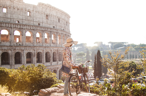 Beautiful young woman in colorful fashion dress walking alone on hill with her bike in front of colosseum in Rome at sunset with trees. Attractive tourist girl with straw hat. Lens Flare.