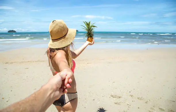 Photo of beautiful girl in swimsuit and pineapple walks on the beach holding the hand of the guy