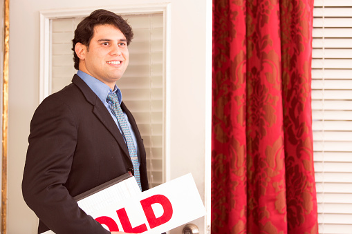 Hispanic real estate agent examines a home he has for sale.  He holds paperwork, portfolio, and Real Estate Agent signs as he enters the home to meet potential buyers.