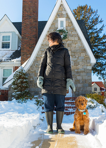 Mature woman with her pet Miniature goldendoodle outdoors in front of their home on a snow covered yard