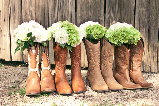 Cowboy boots and bouquets are lined up by a grey barn at a country theme wedding
