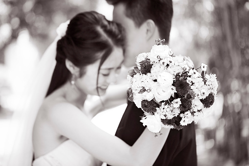 young asian newly wed couple hugging each other during outdoor wedding ceremony, focus on the bouquet, black and white.