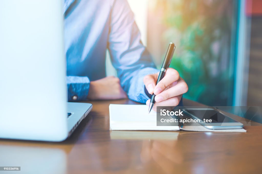 Business woman hand is writing on notepad with pen in office. Business woman hand is writing on notepad with pen in office.On the table are computers, laptops and mobile phones. Book Stock Photo