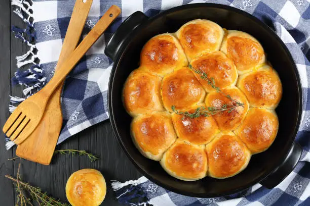 freshly baked soft and fluffy homemade dinner rolls in baking dish with kitchen towel and spatulas on wooden kitchen table, view from above, close-up