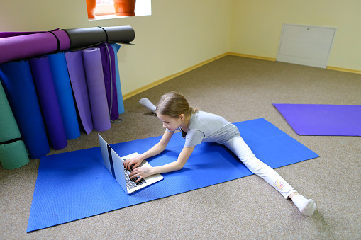 Blonde schoolgirl of European appearance sits on floor in cross-twist position and rewrites with friends through Internet on note. Child focuses on monitor and prints message. Concept of important for children to have healthy lifestyle from early years, or in ability to communicate with loved ones through Internet.