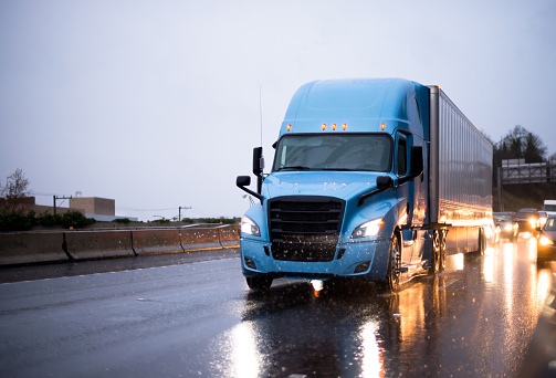 Modern big rig blue semi truck with covered semi trailer driving on twilight rainy wet highway with headlight traffic reflection of the rain drops