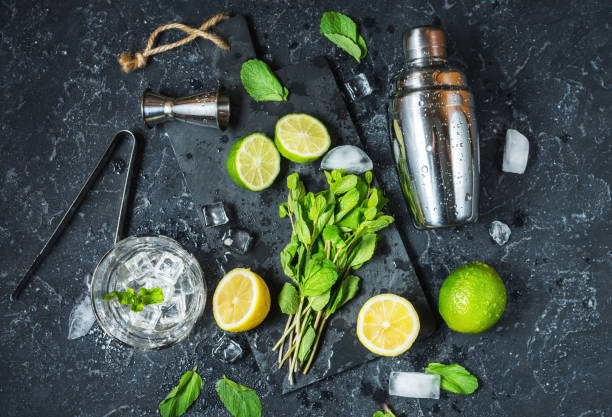 istock Mojito cocktail making. Mint, lime, glass, ice, ingredients and shaker on black stone background. Top view 910348004