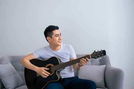 Handsome asian man playing on the guitar on the couch at home