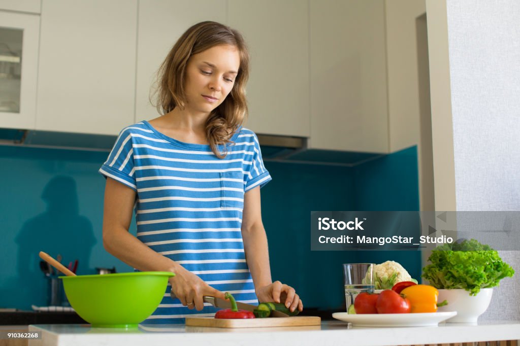 Content Woman Cooking Salad at Kitchen Table Closeup portrait of content young beautiful woman slicing cucumber and cooking salad in kitchen. Healthy cooking concept. Adult Stock Photo