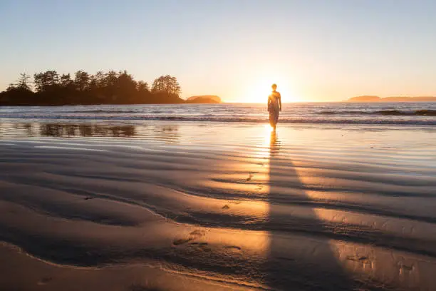 Woman in a casual dress is walking bare feet on a sandy beach during a colorful and vibrant sunset. Taken in Tofino, Vancouver Island, British Columbia, Canada.