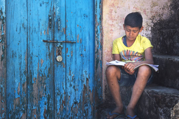 kid reading near locked door Boy sitting on steps near the door with rusty lock and reading, the concept of loneliness india poverty stock pictures, royalty-free photos & images