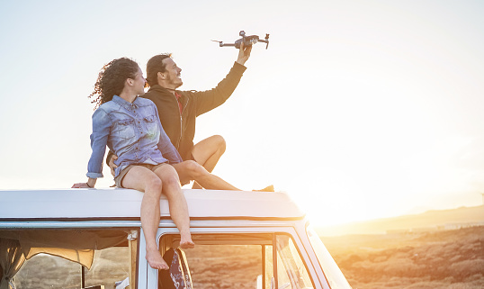 Happy couple using a drone sitting on top of vintage minivan at sunset - Young people having fun with new technology trends during holidays - Travel, tech and vacation concept - Focus on bodies