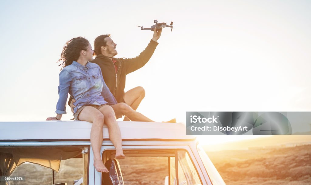 Happy couple using a drone sitting on top of vintage minivan at sunset - Young people having fun with new technology trends during holidays - Travel, tech and vacation concept - Focus on bodies Drone Stock Photo