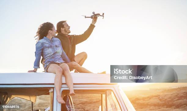 Happy Couple Using A Drone Sitting On Top Of Vintage Minivan At Sunset Young People Having Fun With New Technology Trends During Holidays Travel Tech And Vacation Concept Focus On Bodies Stock Photo - Download Image Now