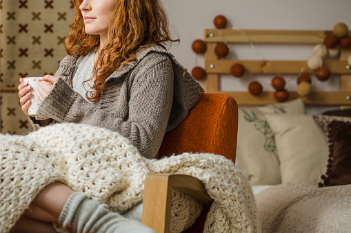 Red-haired woman drinking hot tea while sitting on bed with knit blanket during winter