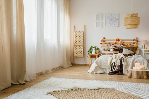 Candles on wood next to bed with knit blanket in hygge style bedroom interior with brown curtain and posters