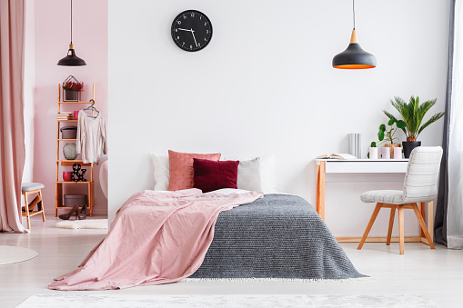 Pink blanket on bed next to desk and grey chair in bedroom interior with black lamp and clock