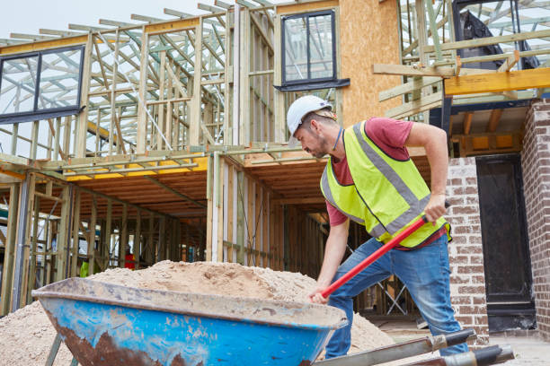 Construction worker shoveling sand by wheelbarrow. Carpenter is working at site. He is in reflective clothing.