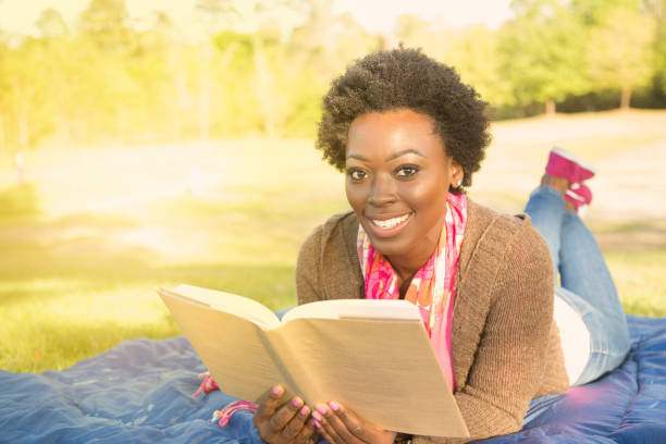 One African descent woman outdoors reading a book in a park during spring or summer season.  The beautiful young adult wears a pink shirt and jacket and lies on a blue blanket.