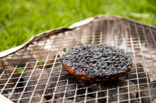 A hamburger barbequing on a grill with charcoal. Low angle perspective with grass in the background.