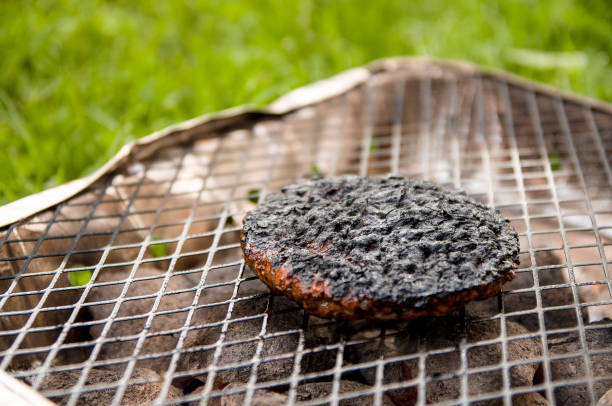 Close-up of a burned burger on a grill in the park. Selective focus. A hamburger barbequing on a grill with charcoal. Low angle perspective with grass in the background. music festival camping summer vacations stock pictures, royalty-free photos & images