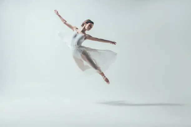 Photo of elegant ballet dancer in white dress jumping in studio