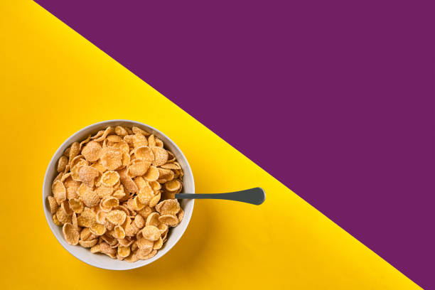 istock Bowl with corn flakes and spoon on purple and yellow background, top view 910620486