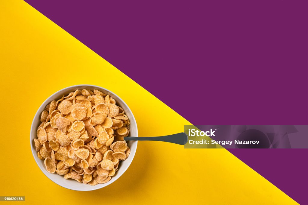 Bowl with corn flakes and spoon on purple and yellow background, top view Food, healthy eating, people and diet concept. Bowl with corn flakes and spoon on purple and yellow background, top view. Copy space. Still life. Flat lay. Cereal Plant Stock Photo