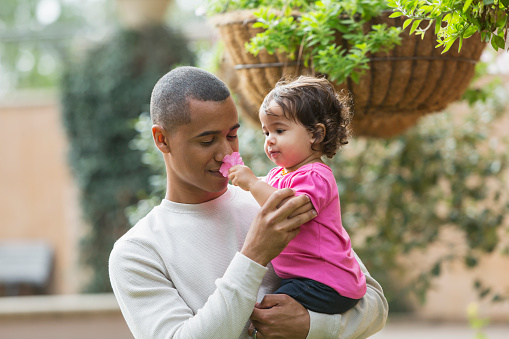 A mixed race young man holding his little girl in his arms. She is holding a flower to his nose.