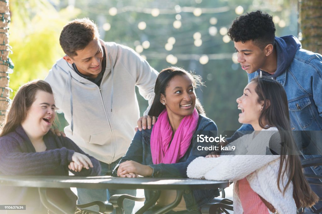 Five teenage friends talking, one with down syndrome A group of five multi-ethnic teenagers sitting and standing around a table outdoors, talking and smiling on a sunny day. The girl sitting on the left has down syndrome. Teenagers Only Stock Photo