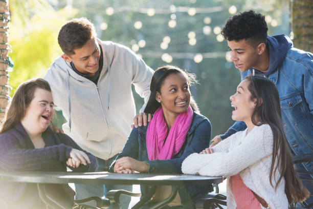 Five teenage friends talking, one with down syndrome A group of five multi-ethnic teenagers sitting and standing around a table outdoors, talking and smiling on a sunny day. The girl sitting on the left has down syndrome. teenagers only stock pictures, royalty-free photos & images