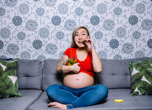 Young pregnant Asian woman sitting on the sofa against wall paper wall in living room. She is wearing jeans and red top with exposed pregnant belly. She is holding a jar of dill pickels and biting one. Her arms  featuring tattoo art . Typical pregnancy cravings.